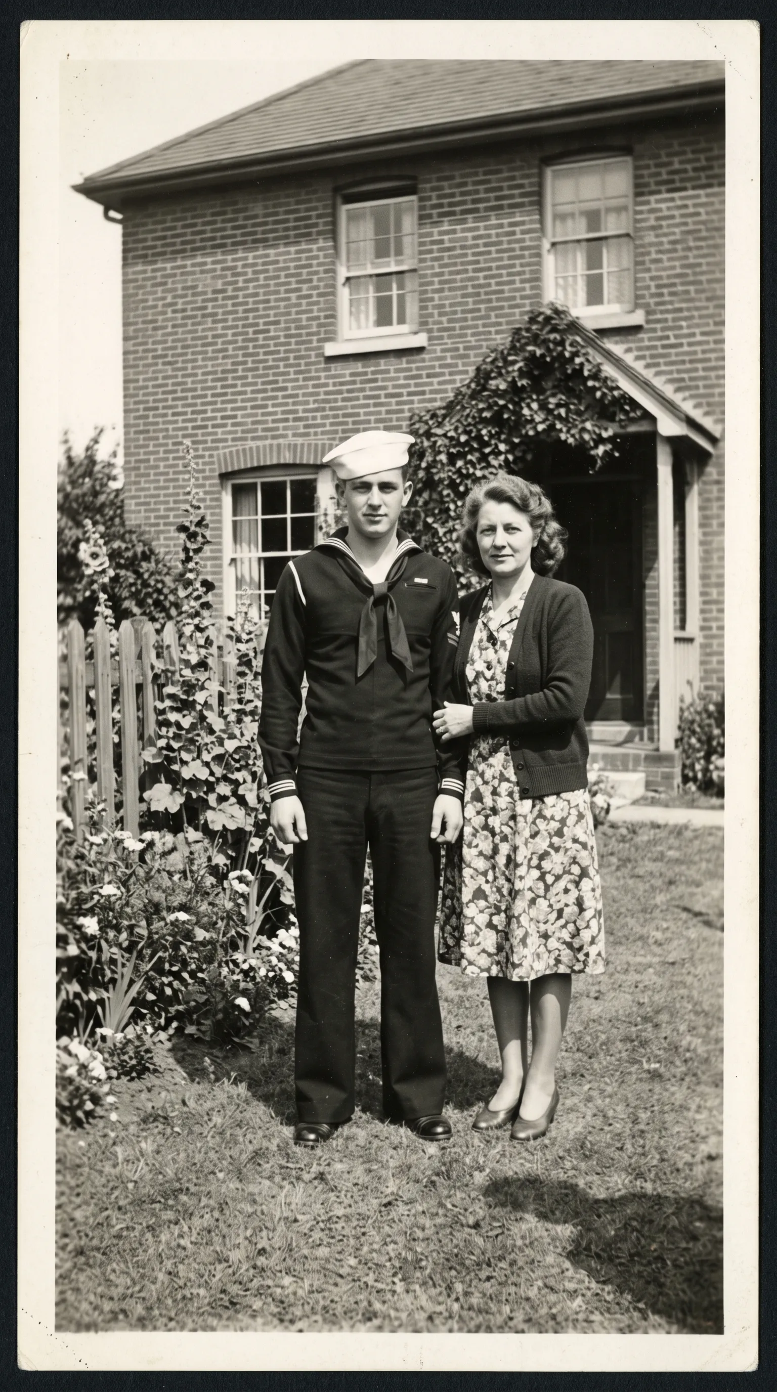 Black-and-white 1940s photo of a young sailor in uniform standing next to his mother outside a brick house