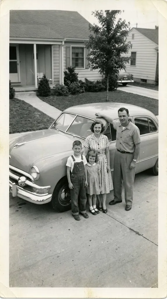 Faded, scratched 1950s photo of a family standing beside their new car outside a house