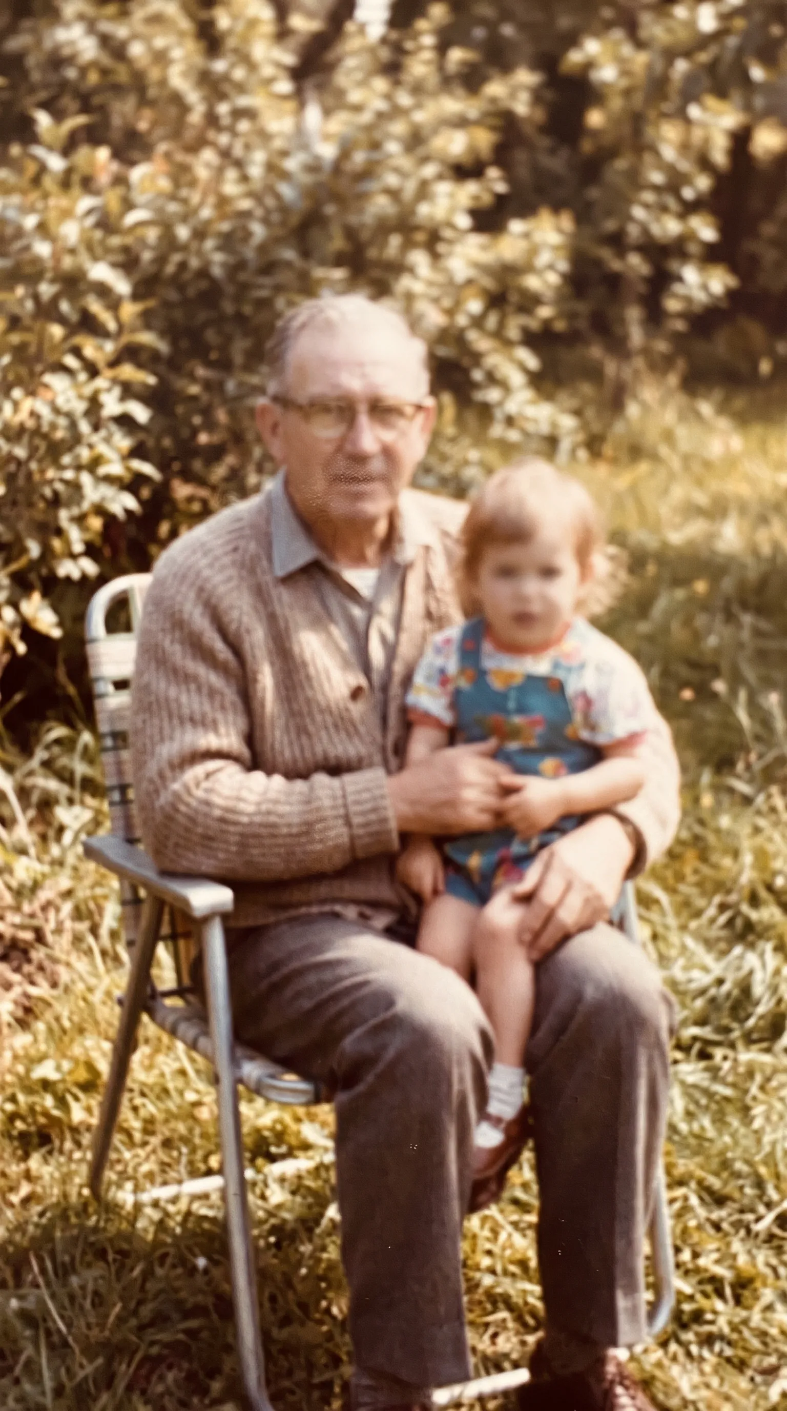 Out-of-focus 1960s color photo of a grandfather holding his granddaughter in a garden